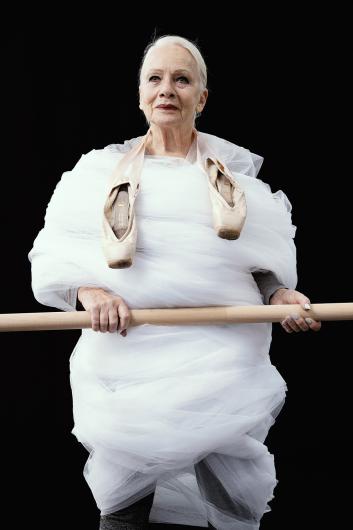 Eine ältere Frau, in weißen Tüll gehüllt, steht selbstbewusst und hält eine Holzstange. Ballettschuhe hängen von ihren Schultern, während sie geradeaus blickt, vor einem schwarzen Hintergrund.