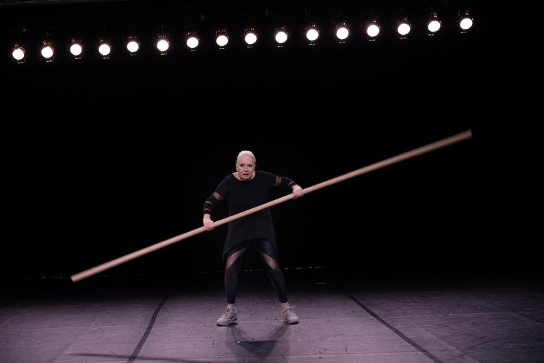 A woman holds a long pole horizontally, bending her knees and leaning forward as if balancing or moving it with force. She has a focused look on her face.
