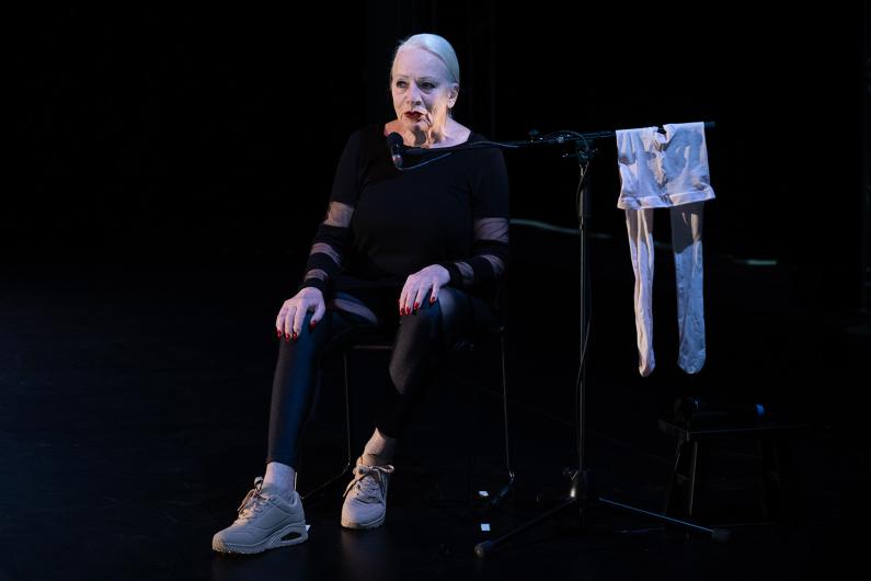A woman sits on a chair with a microphone near her mouth. A pair of white tights is draped over the microphone stand. She wears black leggings and beige sneakers.