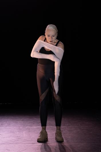A woman slips her arms into a pair of white tights, stretching them across her chest. She wears a black sleeveless top and dark leggings, standing on a stage.