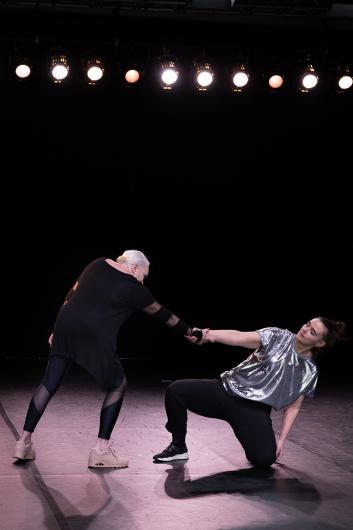 Two performers are on stage: one woman pulls another, who is crouched and wearing a shiny silver shirt, while extending her arm. The background is dark with stage lighting above.