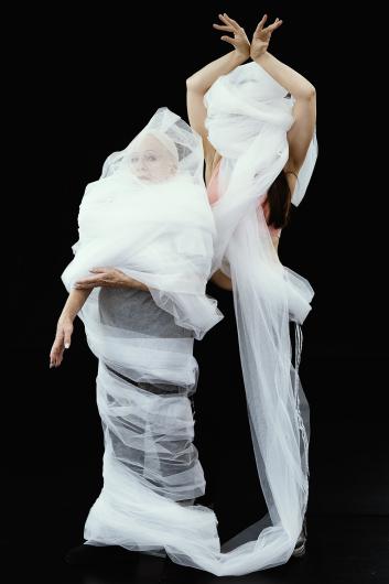 Two individuals wrapped in layers of white tulle stand together against a black background. One person appears older with visible facial features, while the other is partially obscured by the fabric, posing with hands raised in an expressive gesture.