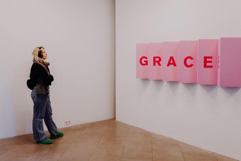 A woman wearing headphones observes an art installation featuring pink panels with the word "GRACE" written in red letters. She stands with her arms crossed, wearing jeans and a scarf.