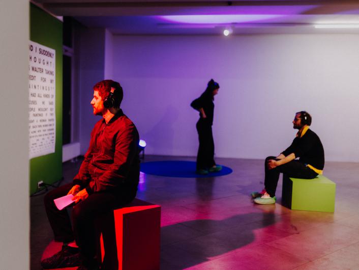 Three people wearing headphones are in a dimly lit room with colorful light reflections. Two of them sit on colored cubes while a third person stands in the background.