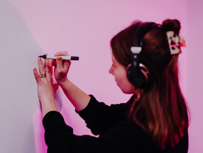 A woman wearing headphones is drawing on a white wall with a marker. Her hand is illuminated by pink light.