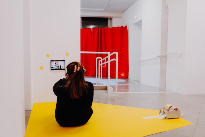 A woman wearing headphones sits on a yellow surface in front of a small screen installation. She looks intently at the display, with a red curtain visible in the background.