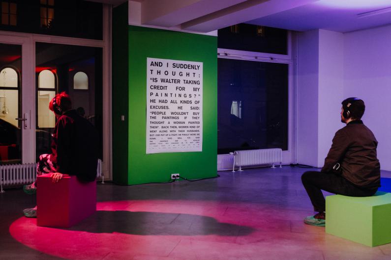 Two people wearing headphones sit on colorful cubes in a dimly lit room, reading a large text on a green wall. The lighting is in shades of purple and red.