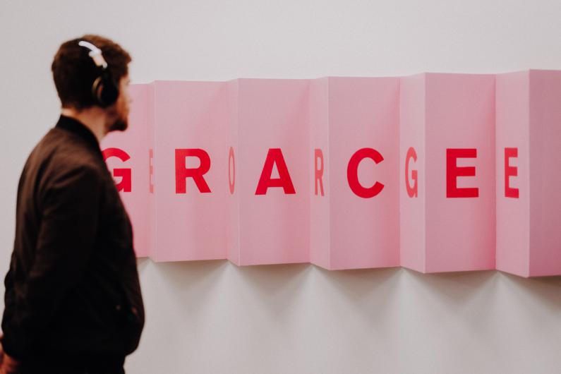 A man wearing headphones observes an art installation made of pink panels. The letters on them form a changing word, depending on the point of view.