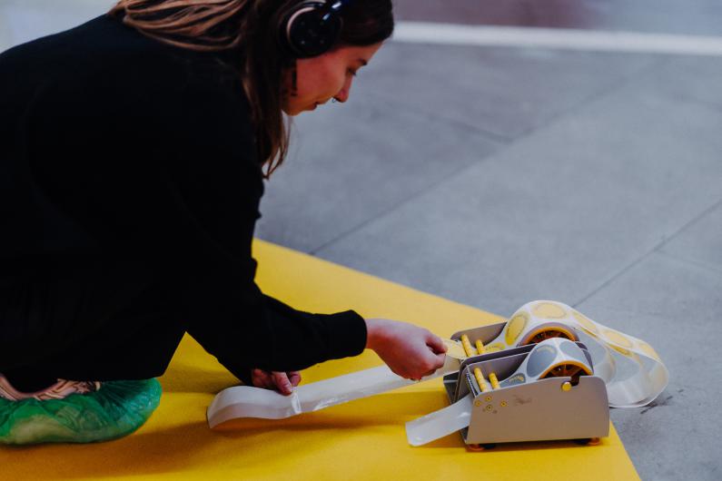 A woman wearing headphones pulls a strip of tape from a special dispenser marked with yellow circles. She is wearing green shoe covers.
