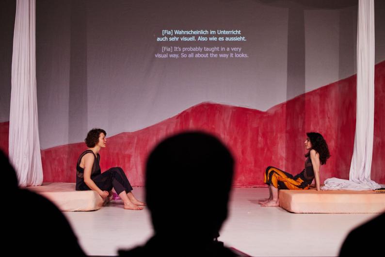 Two performers are sitting on separate cushions on a stage with a red and white backdrop, facing each other. Subtitles are projected above them in both German and English, reading: '[Fia] It's probably taught in a very visual way. So all about the way it looks.' The silhouettes of audience members are visible in the foreground.