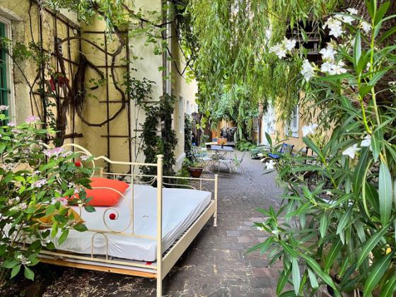 A picturesque, green courtyard featuring a white metal bed with a mattress, a white sheet, and a decorative pillow with an orange accent. The surroundings are filled with climbing plants, blooming shrubs, and hanging branches, creating a peaceful, almost fairytale-like atmosphere. In the background, additional seating and tables invite relaxation.
