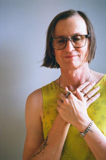 A portrait of a person with medium-length brown hair and glasses, gently smiling with closed eyes. They are wearing a sleeveless yellow-green top with floral patterns and have both hands crossed over their chest. They wear multiple rings, a bracelet, and a necklace. The image has warm, soft lighting against a simple, neutral background.