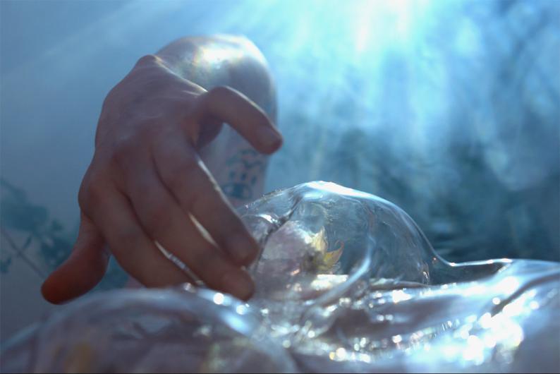 A close-up of a hand touching a transparent, organically shaped surface. Sunlight streams through the scene, creating a dreamy, almost otherworldly atmosphere.