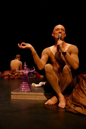Eine performende Person sitzt auf der Bühne, hält ein Mikrofon und macht eine mudra-artige Geste mit der Hand. Daneben steht eine violette Skulptur in Frauenform.