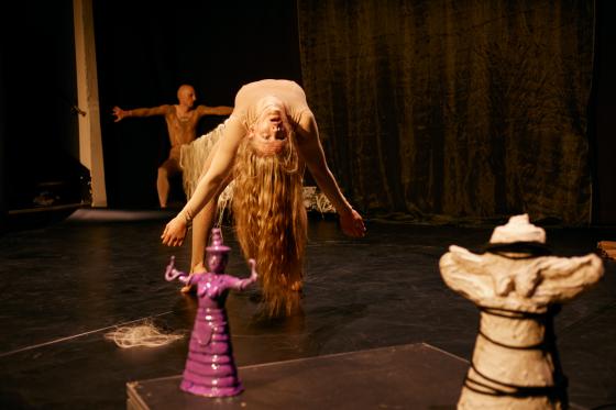 A performer leans dramatically backward, their long hair cascading to the floor. A purple statue is visible in the foreground.