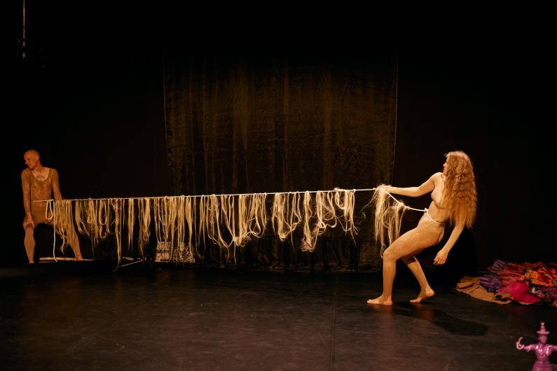 Zwei performende Personen ziehen an einem langen Seil aus hellem Kunsthaar über die Bühne, was wie ein symbolischer Kraftakt wirkt.