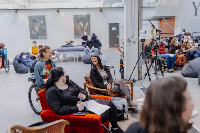 A modern, open event or conference hall with people sitting comfortably in armchairs and wheelchairs. A woman in the center is smiling and engaging in conversation with another person. In the background, more attendees are sitting on bean bags and chairs, some wearing masks. Cameras and recording equipment are set up to document the event.