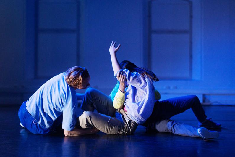 Three dancers in a contemporary performance on a dimly lit stage with blue lighting. They are intertwined on the floor, with dynamic body positions and expressive gestures.