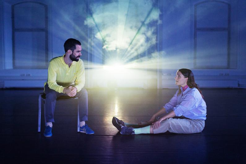 A man in a yellow sweater sits on a chair, looking at a woman sitting on the floor with her legs stretched out. A projection with radiant lights illuminates the background.