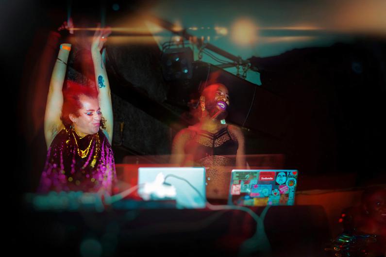 A photo of two DJs or performers standing at their laptops on a stage. The person on the left wears a shimmering purple outfit and raises their arms in celebration. The person on the right wears a black, sheer top and smiles brightly. Colorful lights and motion blur create a dynamic, energetic atmosphere.
