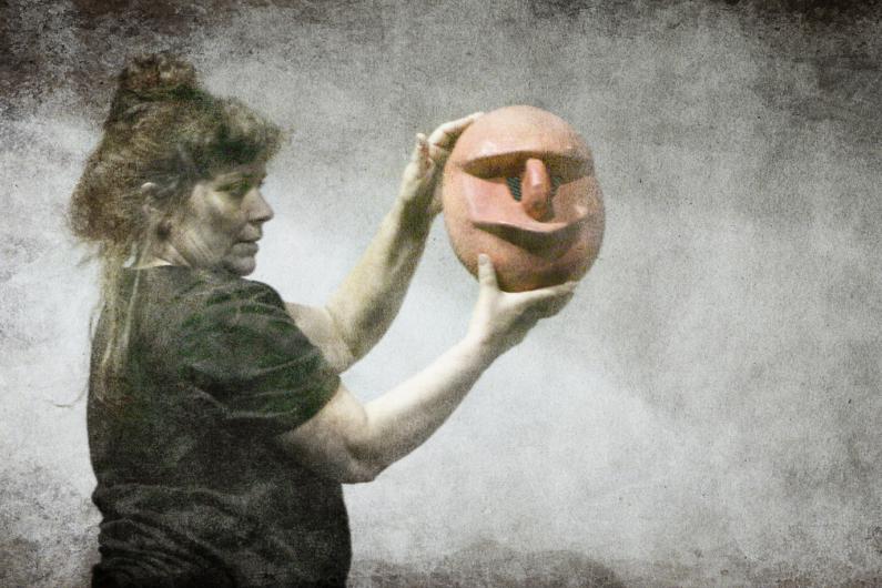 A woman with dark, tied-up hair and a black top holds a large red theatrical mask with a stylized facial expression in both hands. The background is textured and appears foggy, giving the image a mystical or theatrical atmosphere.