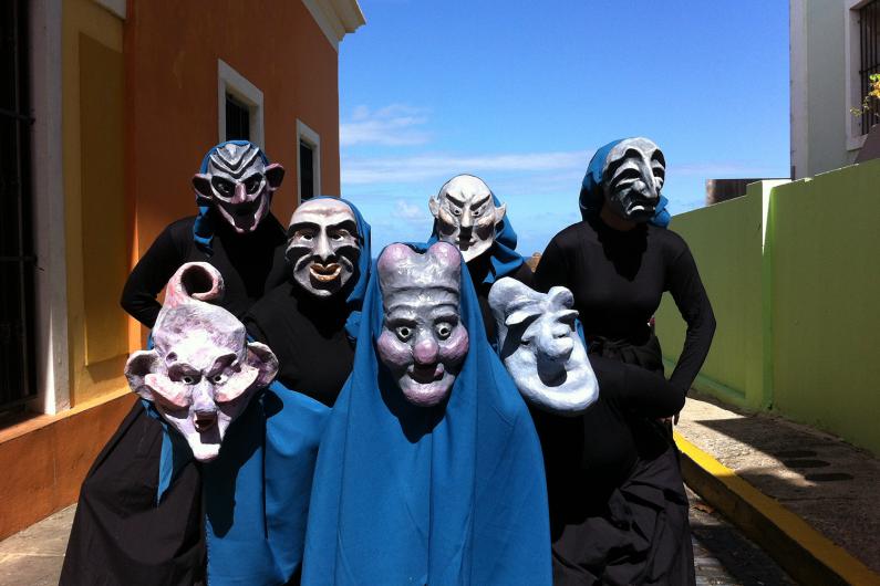 Eine Gruppe von sechs Personen trägt große, expressive Masken mit verschiedenen Gesichtsausdrücken. Sie sind in schwarze Kleidung und blaue Tücher gehüllt. Die Szene spielt sich im Freien vor einer farbenfrohen Häuserfassade und einem strahlend blauen Himmel ab.