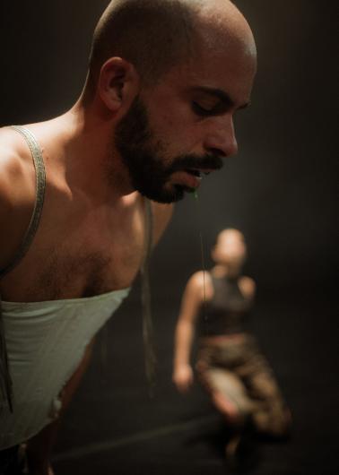 A bald man with a beard leans forward as a thin green strand of saliva drips from his mouth. He is wearing a tight, light-colored top with straps. In the blurred background, a person is kneeling on the floor.