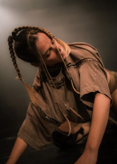 A performer with braided hair moves on the floor. She wears a loose, beige top with decorative details. Her head is slightly tilted, and her gaze is directed into the distance.