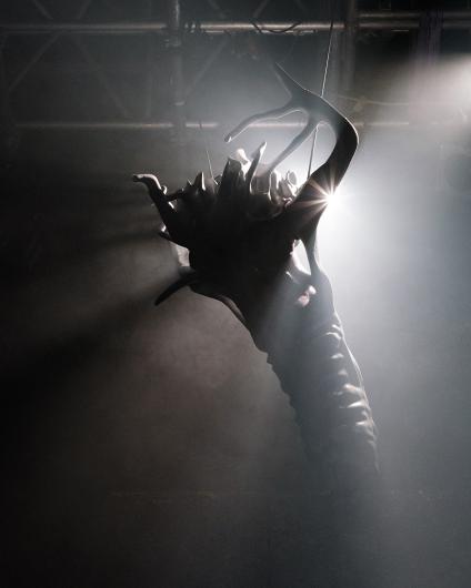 A sculptural hand with branched, organic shapes rises from the darkness. A beam of light hits the tip of the hand, creating dramatic illumination.