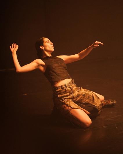 A performer kneels on the floor with her arms outstretched. She wears a black top and a skirt with lace-up details. The scene is bathed in warm, golden light.