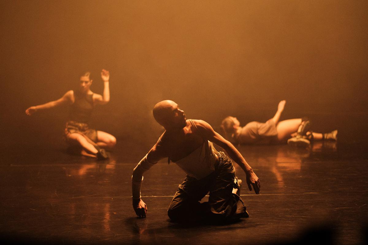 Three performers are on a stage enveloped in warm, golden light. A man in the center kneels with his head tilted, while two dancers in the background take different poses.