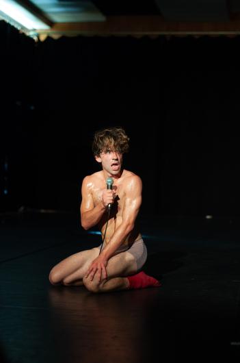 A muscular dancer kneels on a stage, holding a microphone. His upper body is covered in sweat, and his facial expression is intense. He is wearing tight underwear and red socks. The stage lighting makes him stand out against the dark background.