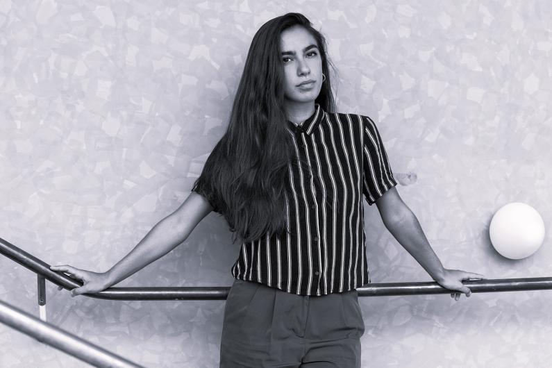 A young woman with long dark hair leans against a railing. She wears a striped shirt and looks confidently into the camera. The background is a textured wall.