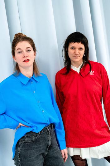 Zwei Frauen stehen vor einem hellblauen, wellenförmigen Vorhang und blicken in die Kamera. Die Frau links trägt eine leuchtend blaue Bluse und dunkle Jeans, ihre Lippen sind rot geschminkt. Die Frau rechts trägt einen roten Adidas-Pullover mit einem weißen Rock und hat dunkles, schulterlanges Haar mit Pony. Beide wirken selbstbewusst und entspannt.