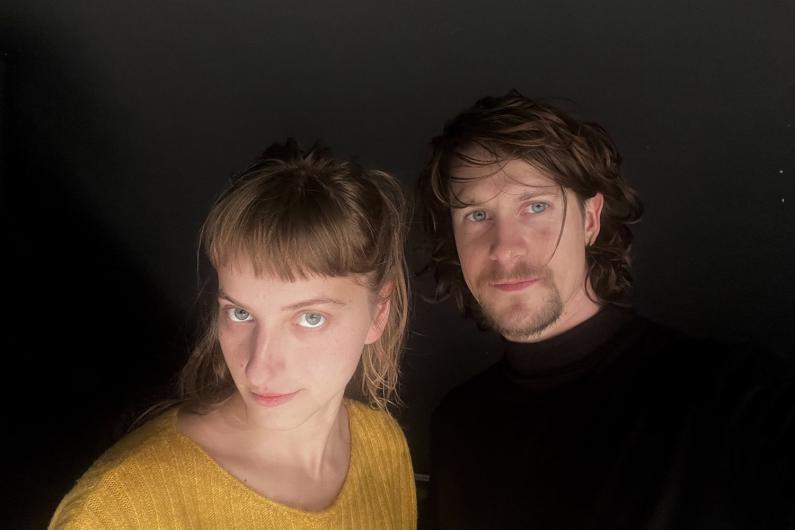 A man and a woman pose for a selfie against a dark background. The woman wears a yellow sweater and has bangs, while the man has curly hair and a black sweater.
