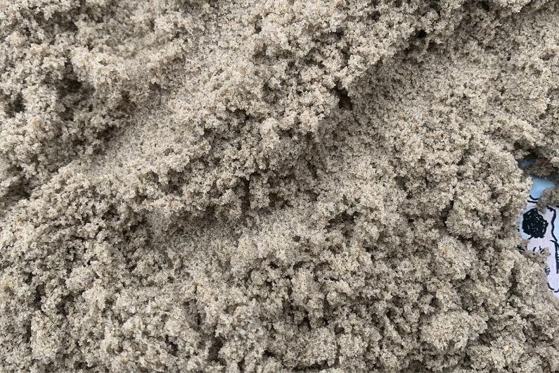 Eine Nahaufnahme von lockerem Sand mit einer groben, unregelmäßigen Struktur. Die Körner sind deutlich sichtbar. In der rechten Ecke sieht man die Zeichnung eines Menschen aus dem Sand ragen.