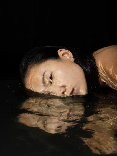 Eine Frau liegt mit ihrem Gesicht halb im Wasser, ihr Blick ist ruhig und nachdenklich. Die Wasseroberfläche erzeugt eine verzerrte Spiegelung ihres Gesichts.