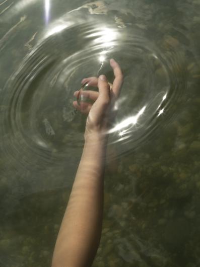 A hand is dipped into water, surrounded by gentle ripples and shimmering light. The reflections on the water’s surface give the image a dreamy aesthetic.