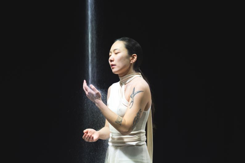 A woman in a flowing white outfit stands under a vertical beam of salt, gazing at her hand with a serene expression.