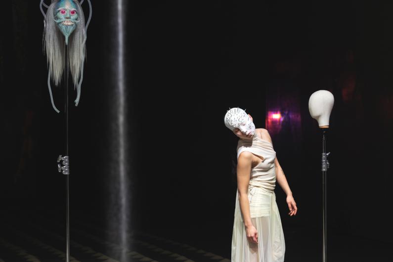 Eine Performerin mit einer stacheligen weißen Maske neigt sich zur Seite zwischen zwei auf Ständern montierten Schaufensterpuppenköpfen in einem dunklen Raum.