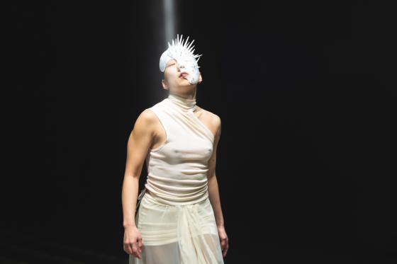 Eine Figur in einem cremefarbenen, einseitig schulterfreien Outfit blickt nach oben, während ein Scheinwerfer eine stachelige Maske auf ihrem Gesicht beleuchtet.