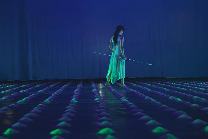 Eine barfüßige Performerin in einem transparenten Kleid geht unter blauem Licht mit einer langen Stange über ein Raster aus kleinen Reishaufen.