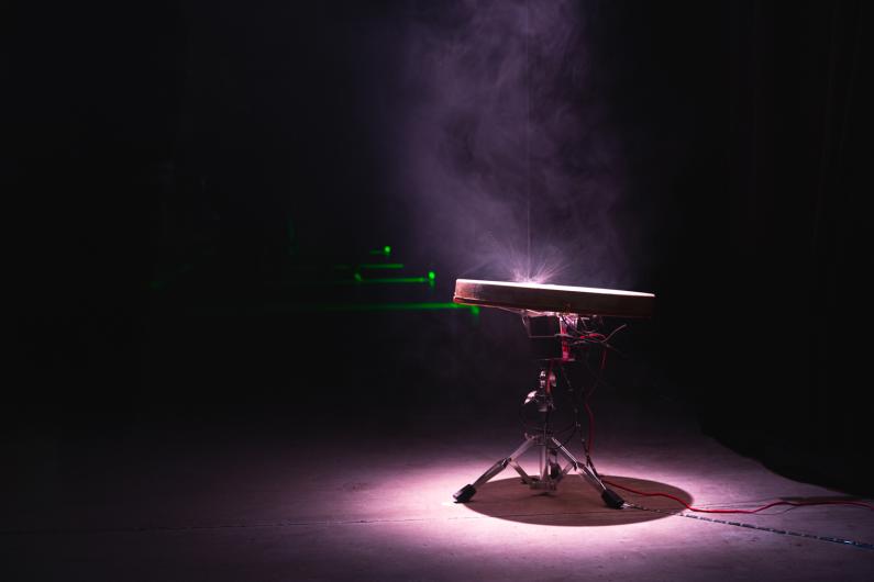 A circular drum emits light and smoke on a stage, surrounded by cables and electronics in a dark setting. Water drips from above onto the drum.