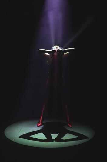 Eine Performerin in einem roten Outfit steht im Scheinwerferlicht, den Kopf zurückgelehnt, während Licht aus einer dramatischen Maske strömt.