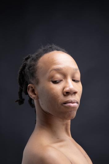 Ein Porträt einer Person mit geflochtenen Haaren vor einem schwarzen Hintergrund. Die Person hat einen ruhigen und introspektiven Gesichtsausdruck, mit leicht gesenktem Blick und geschlossenen Lippen.