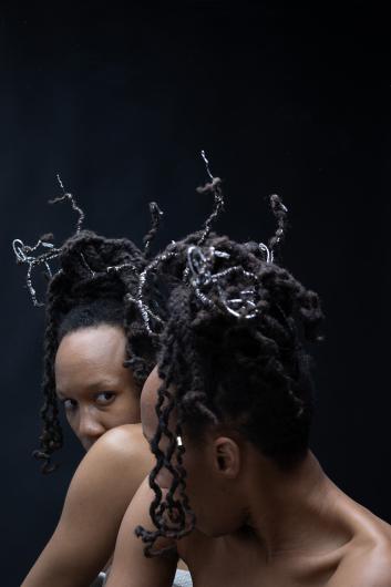 A person sits in front of a mirror, looking over her shoulder towards the camera. Their hair is adorned with metal spirals protruding from the curls. The reflection in the mirror enhances the mysterious and contemplative mood.