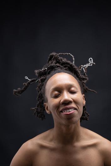 Ein Nahaufnahme-Porträt einer lächelnden Person mit geschlossenen Augen. Ihr Haar ist mit Drahtspiralen verziert, die sich kunstvoll um die Locken winden. Der dunkle Hintergrund hebt das strahlende Gesicht hervor.