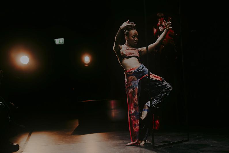 A dancer elegantly balances on one leg with raised arms, holding onto a metal structure, as two warm lights glow in the background.