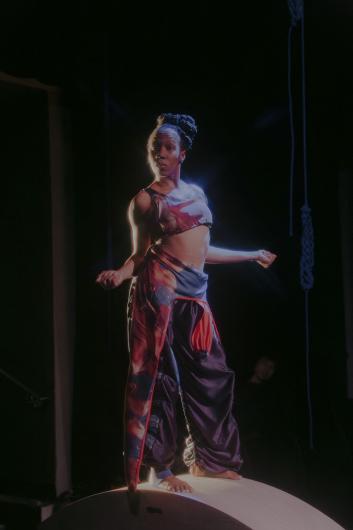 A dancer with an updo stands on a curved platform, illuminated by cool lighting, her outfit featuring a red-blue pattern.