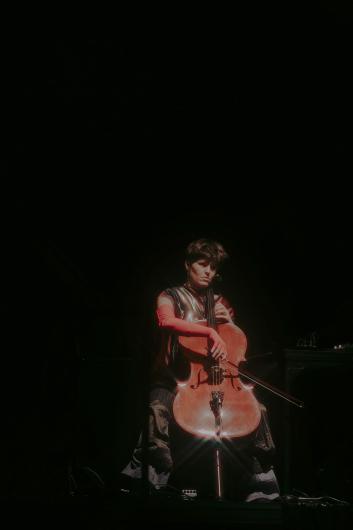 A cellist sits in a dark space, playing their instrument with a thoughtful expression as a soft glow highlights the cello.